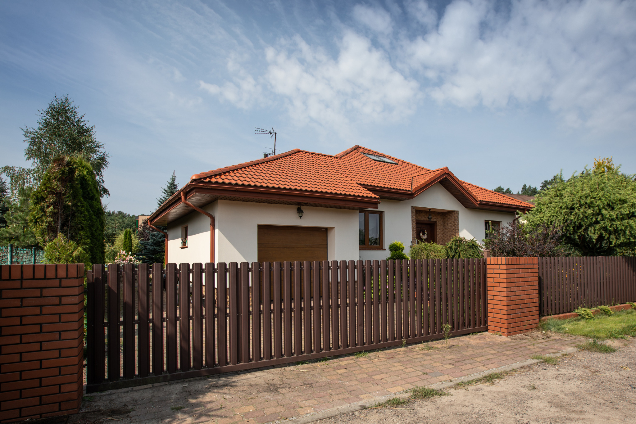 Jednorodzinny dom z czerwoną dachówką, białymi ścianami i brązowym ogrodzeniem. Widok z zewnątrz w słoneczny dzień.