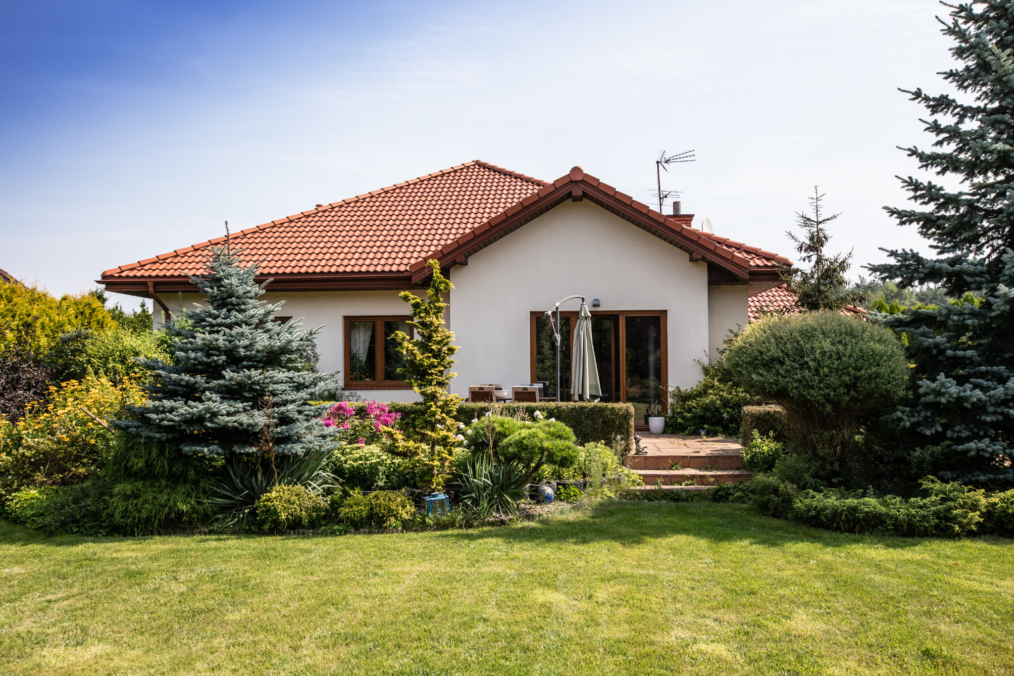 Biały dom z czerwoną dachówką otoczony zielenią, z widocznym tarasem i zadbanym ogrodem. Uwagę przyciąga bujna roślinność i spokojna atmosfera.