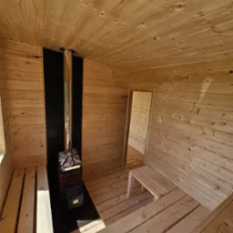 Drewniana sauna z piecem na kamienie w Gostkowie. Jasne drewno, czarny panel za piecem. Wnętrze oświetlone, widoczne ławki i otwór drzwiowy.
