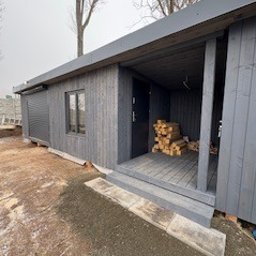 Abies.c.s.s Radosław Papaj - Szara, drewniana altana z podestem i stosem drewna wewnątrz. Widoczne rolety zewnętrzne i drzwi wejściowe. Całość w surowym, minimalistycznym stylu.