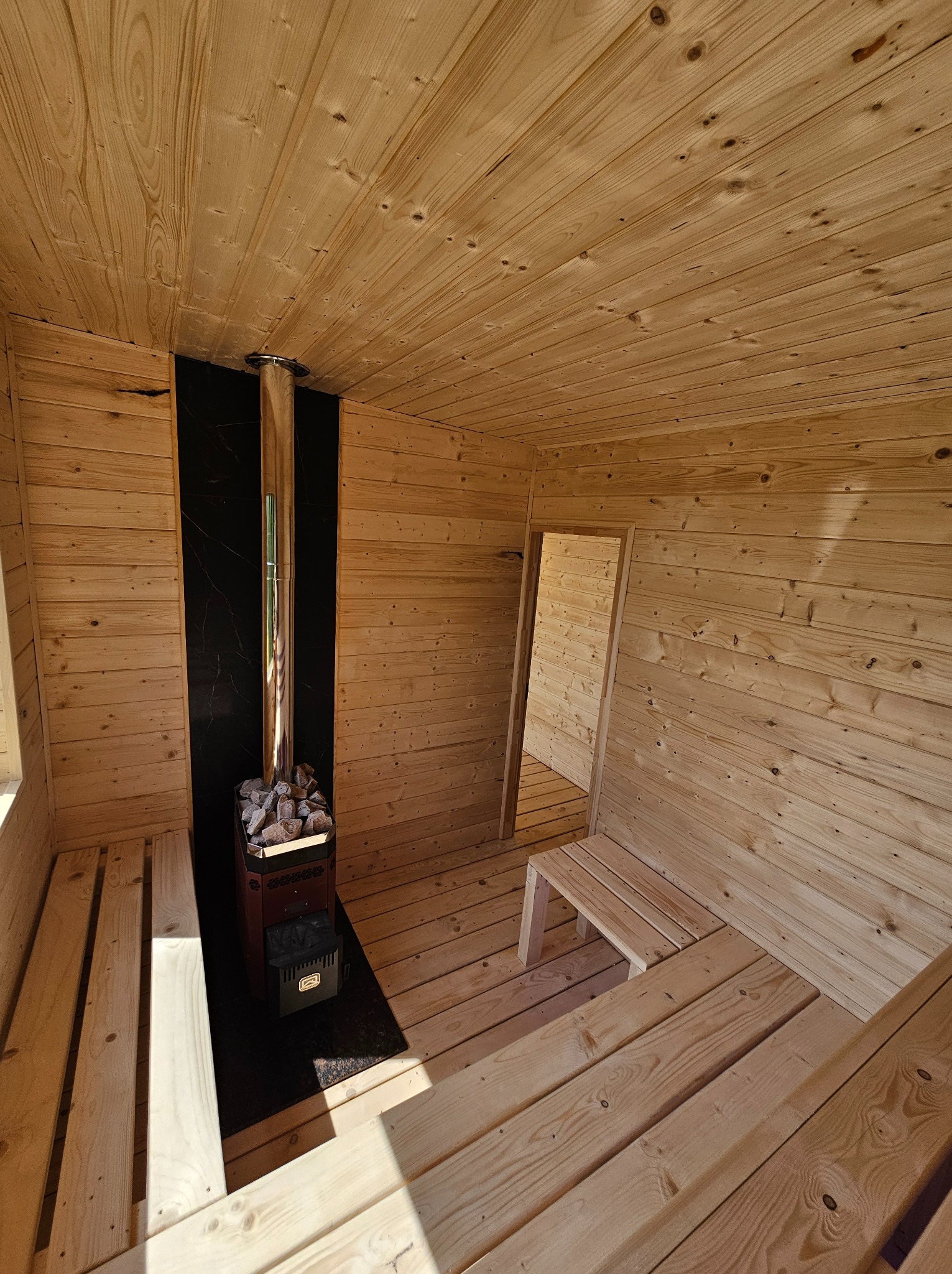 Drewniana sauna z piecem na kamienie w Gostkowie. Jasne drewno, czarny panel za piecem. Wnętrze oświetlone, widoczne ławki i otwór drzwiowy.