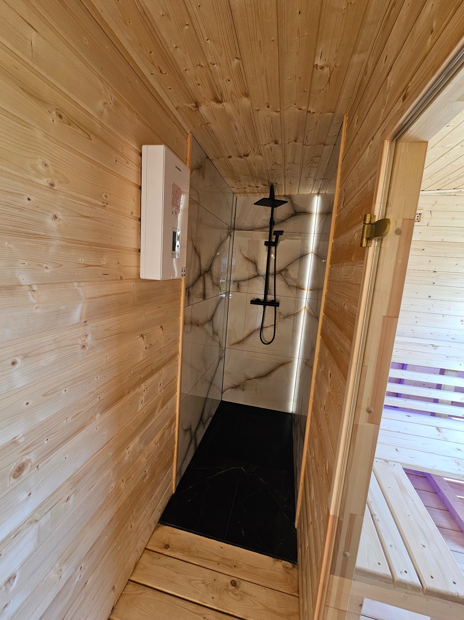 Wnętrze sauny z drewnianymi ścianami, czarną podłogą i kabiną prysznicową z marmurowymi płytkami. Widoczny bojler na ścianie i fragment drewnianej ławki.