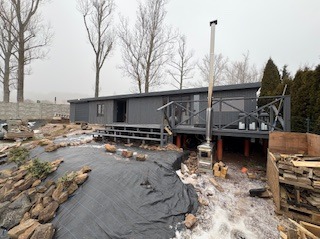 Szara altana z tarasem w Gostkowie, widok z dołu. Elementy wykończeniowe w trakcie prac, teren wokół w trakcie zagospodarowywania. Widoczne drzewa w tle.