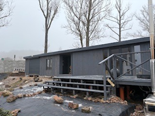 Szara, drewniana sauna z tarasem na nierównym terenie. Widoczne schody, komin i kamienie dekoracyjne. Pochmurne niebo w tle.