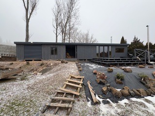 Nowoczesna, szara altana z drewna z dużymi oknami i tarasem, otoczona ogrodem z elementami skalnymi i schodami. Zimowa sceneria z resztkami śniegu.