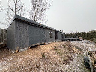 Nowoczesna, grafitowa altana z roletami zewnętrznymi i drewnianym tarasem na nierównym terenie. Widoczne drzewa iglaste w tle i stos drewna.