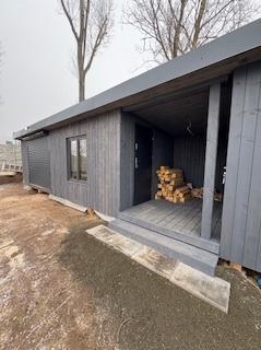 Szara, drewniana altana z podestem i stosem drewna wewnątrz. Widoczne rolety zewnętrzne i drzwi wejściowe. Całość w surowym, minimalistycznym stylu.