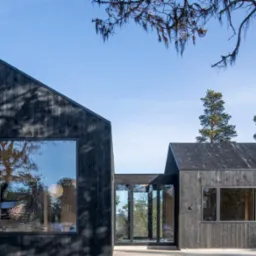 Nowoczesny, czarny budynek z dużymi oknami i drewnianą elewacją, otoczony zielenią i błękitnym niebem, prezentujący minimalistyczny design i architekturę.