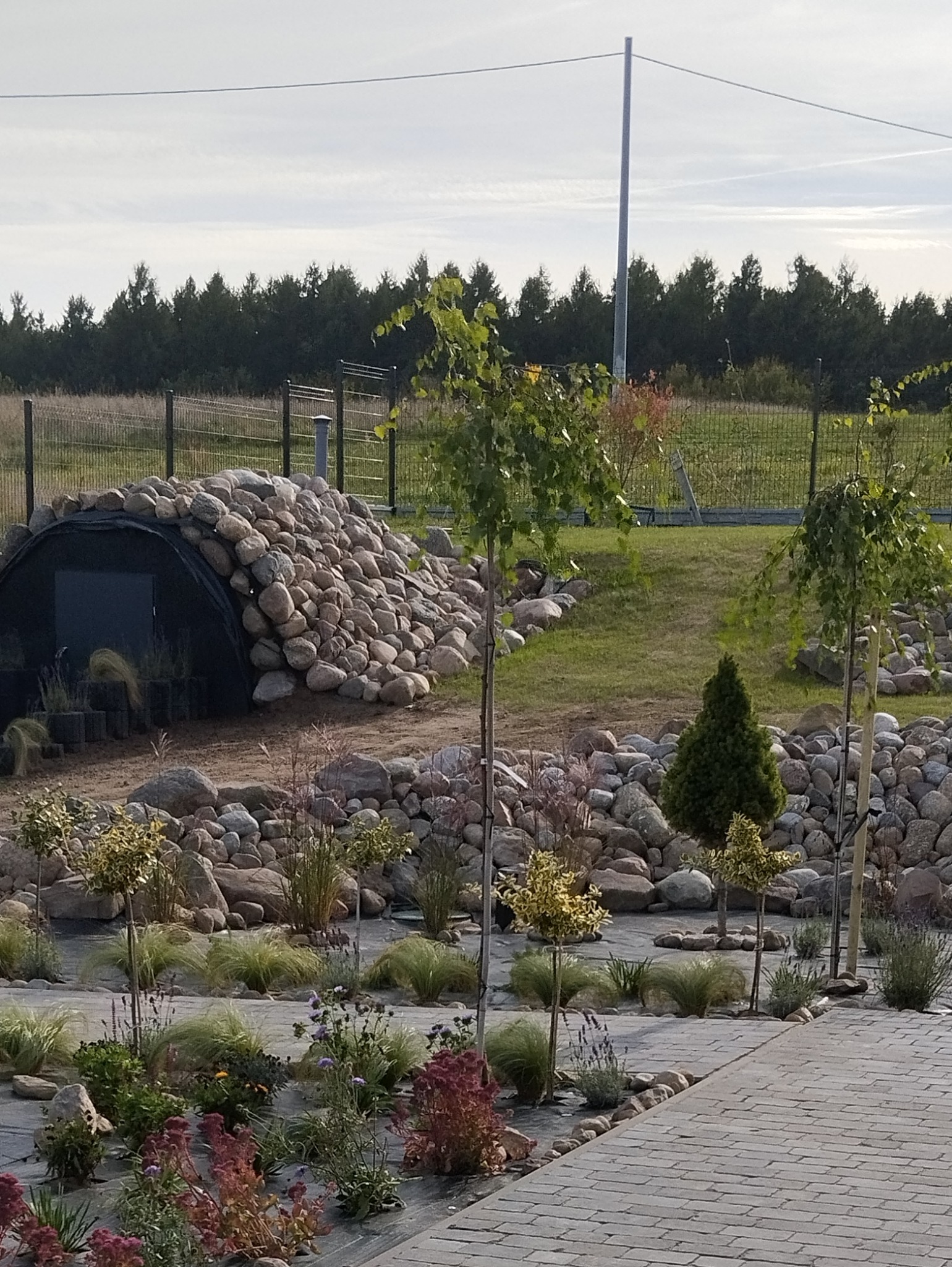 Aranżacja ogrodu z tunelem obsypanym kamieniami, niskimi drzewkami i kwitnącymi rabatami. W tle widoczny las i ogrodzenie.
