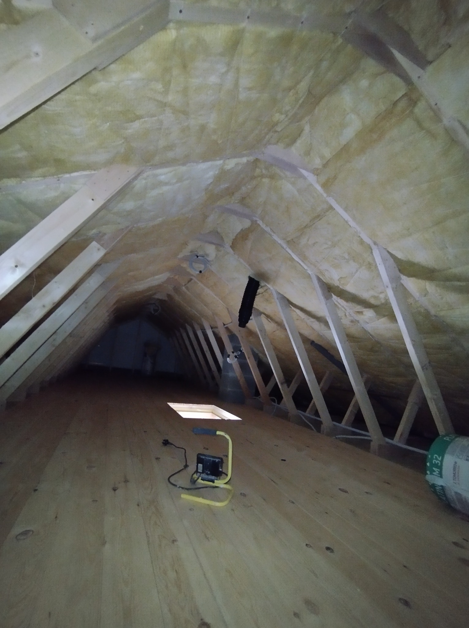 Izolacja poddasza wełną mineralną, widoczne drewniane elementy konstrukcyjne, oświetlenie robocze i rolka materiału izolacyjnego. Ukończony etap prac.