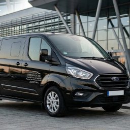 SPEED BLACK TAXI DENYS TYSHYK - Czarny, lśniący bus Ford Transit Custom z logo firmy Speed Black Taxi na tle nowoczesnego budynku lotniska i flagi Polski. Usługi przewozu osób.