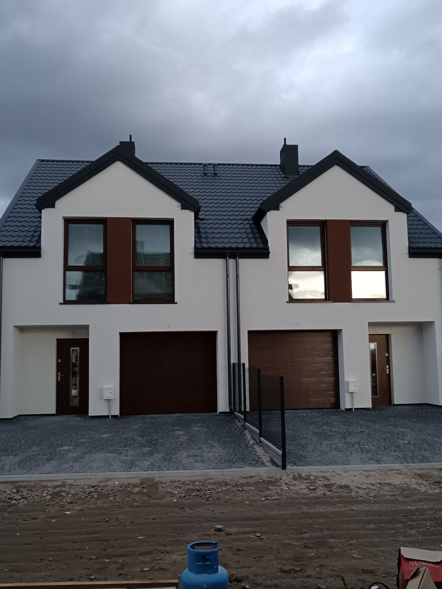 Bliźniak w nowoczesnym stylu z białymi ścianami, brązowymi akcentami okiennymi i bramami garażowymi, pod ciemnym, pochmurnym niebem. Kostka brukowa przed budynkiem.