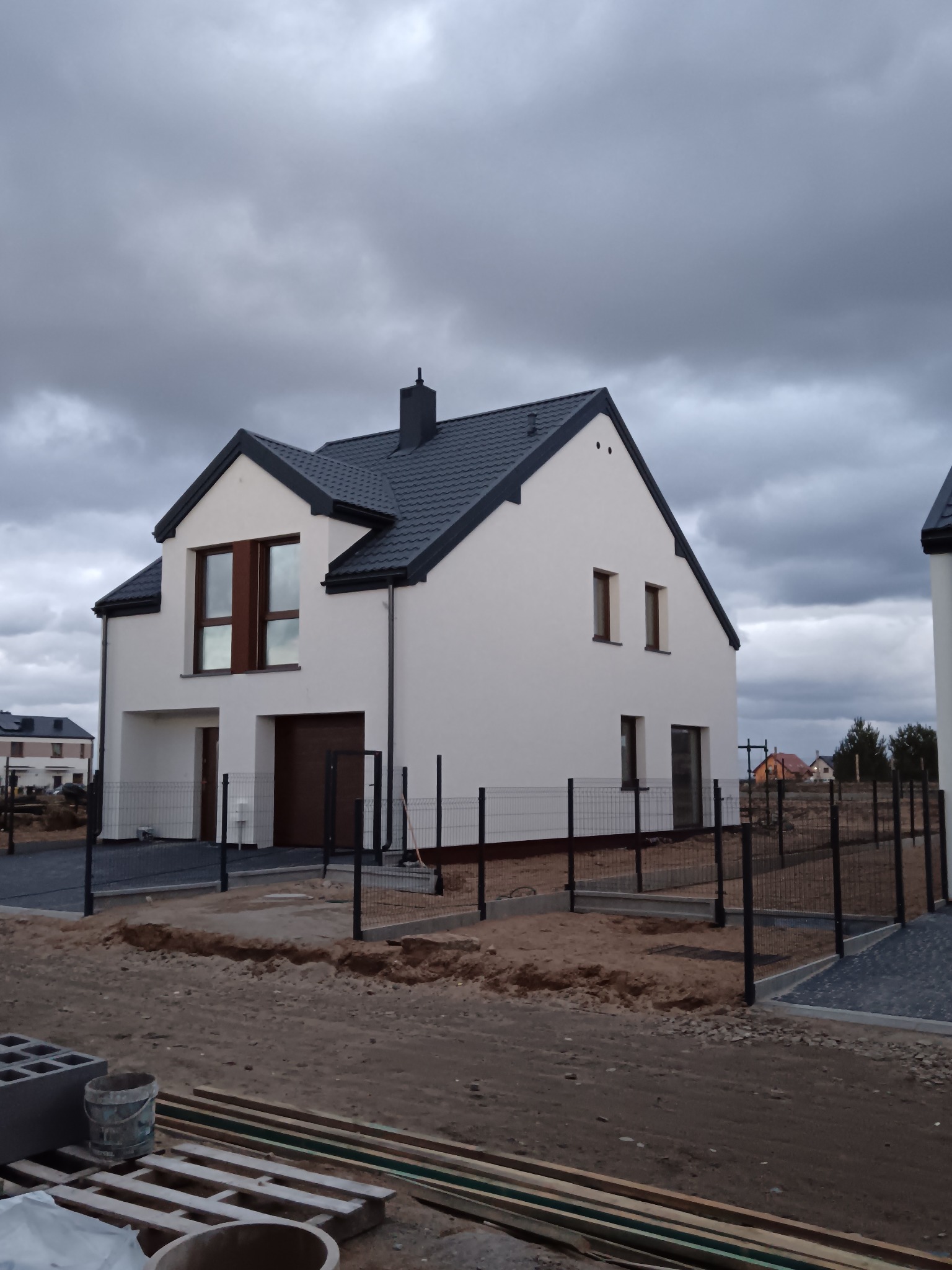 Nowoczesny, dwukondygnacyjny dom z białymi ścianami i ciemną dachówką, otoczony tymczasowym ogrodzeniem. Widoczne materiały budowlane sugerują trwające prace wykończeniowe.
