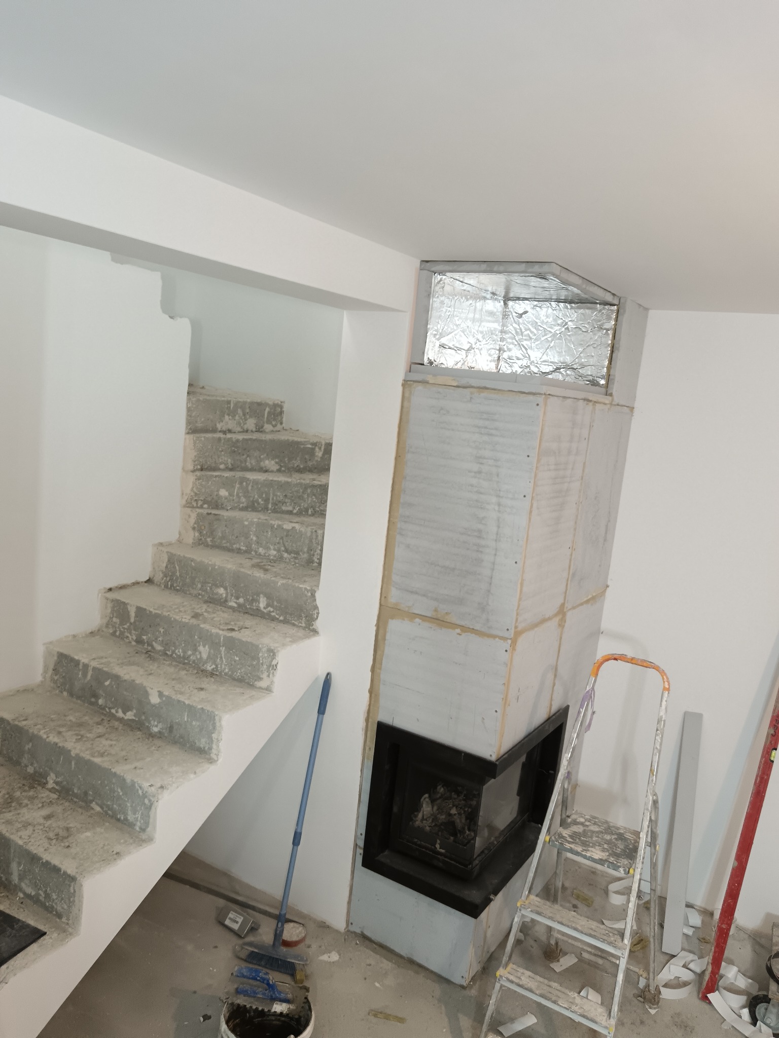 Wnętrze w trakcie wykańczania: surowe schody betonowe, obudowa kominka z płyt gipsowych z folią aluminiową, drabina malarska i narzędzia malarskie na podłodze.