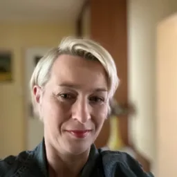 Portret kobiety z krótkimi blond włosami, w dżinsowej kurtce, uśmiechającej się do kamery na rozmytym tle wnętrza.