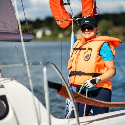 KinderParty MARTA MARCINOW - Uśmiechnięta kobieta w okularach i kapoku na jachcie, trzyma linę. W tle woda i zieleń. Słoneczny dzień, morski klimat, widok z bliska.