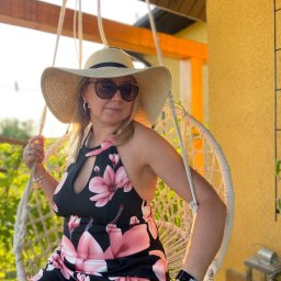 KinderParty MARTA MARCINOW - Kobieta w kapeluszu i okularach przeciwsłonecznych siedzi w białym, wiszącym fotelu na tle zieleni i żółtej ściany z pomarańczowymi elementami.