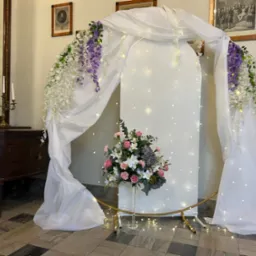 Dekoracja ślubna: biały łuk z kwiatami w odcieniach fioletu i bieli, oświetlony delikatnymi lampkami, z bukietem w szklanym wazonie. Klasyczne wnętrze z obrazami i komodą.