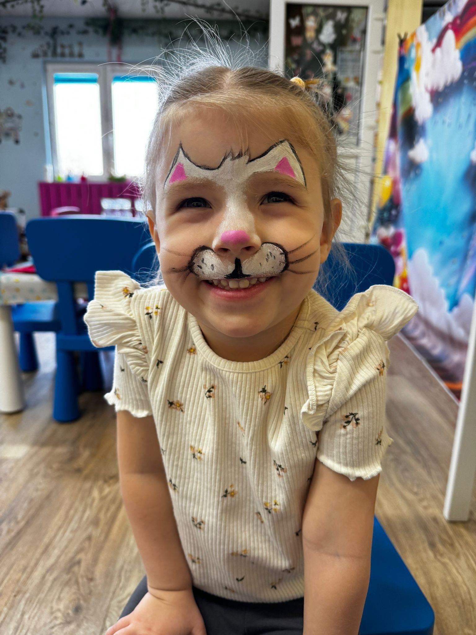 Uśmiechnięta dziewczynka z pomalowaną twarzą w motywy kota, siedząca na niebieskim krześle, w tle pokój dziecięcy z dekoracjami. Zabawa w charakteryzację.