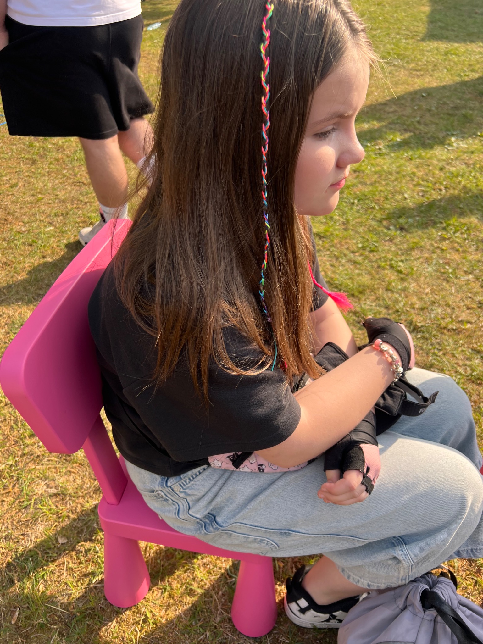 Dziewczynka z warkoczykiem siedzi na różowym krześle na trawie. Ubrana w jeansy i czarną bluzkę, ma na rękach ochraniacze. W tle widać fragmenty innych osób.