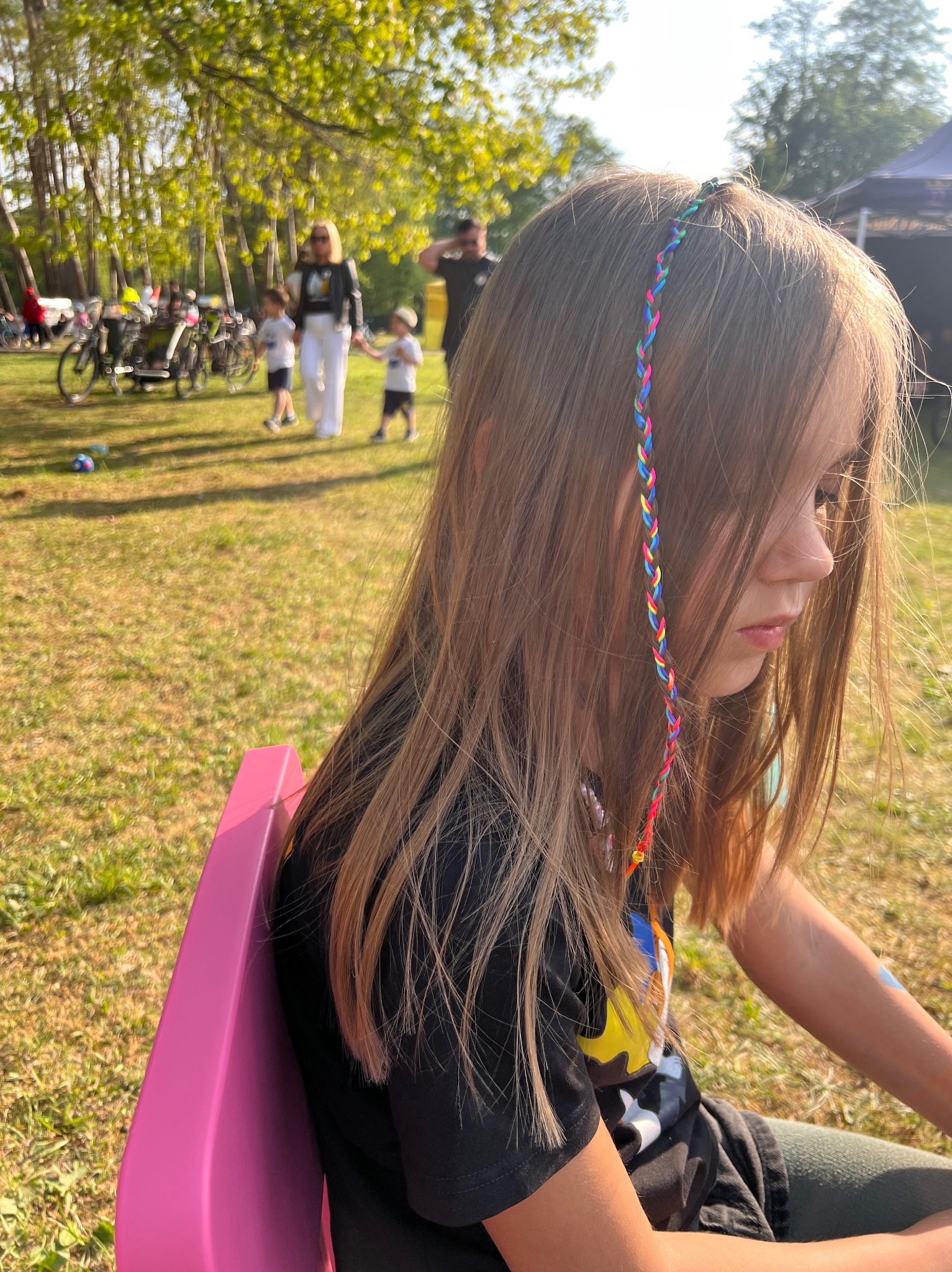 Dziewczynka siedzi na różowym krześle w plenerze, z kolorowym warkoczykiem we włosach. W tle widoczni ludzie i rowery, co sugeruje piknik lub wydarzenie na świeżym powietrzu.