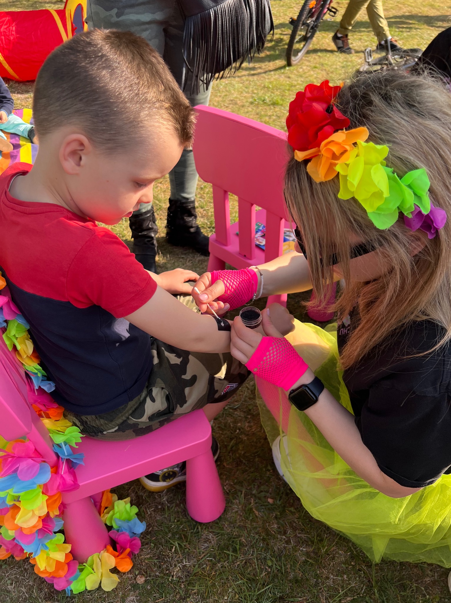 Animator maluje brokatowy tatuaż na ramieniu chłopca siedzącego na różowym krześle, z kolorowymi kwiatami. Artystka w neonowych rękawiczkach i kwiatach we włosach.