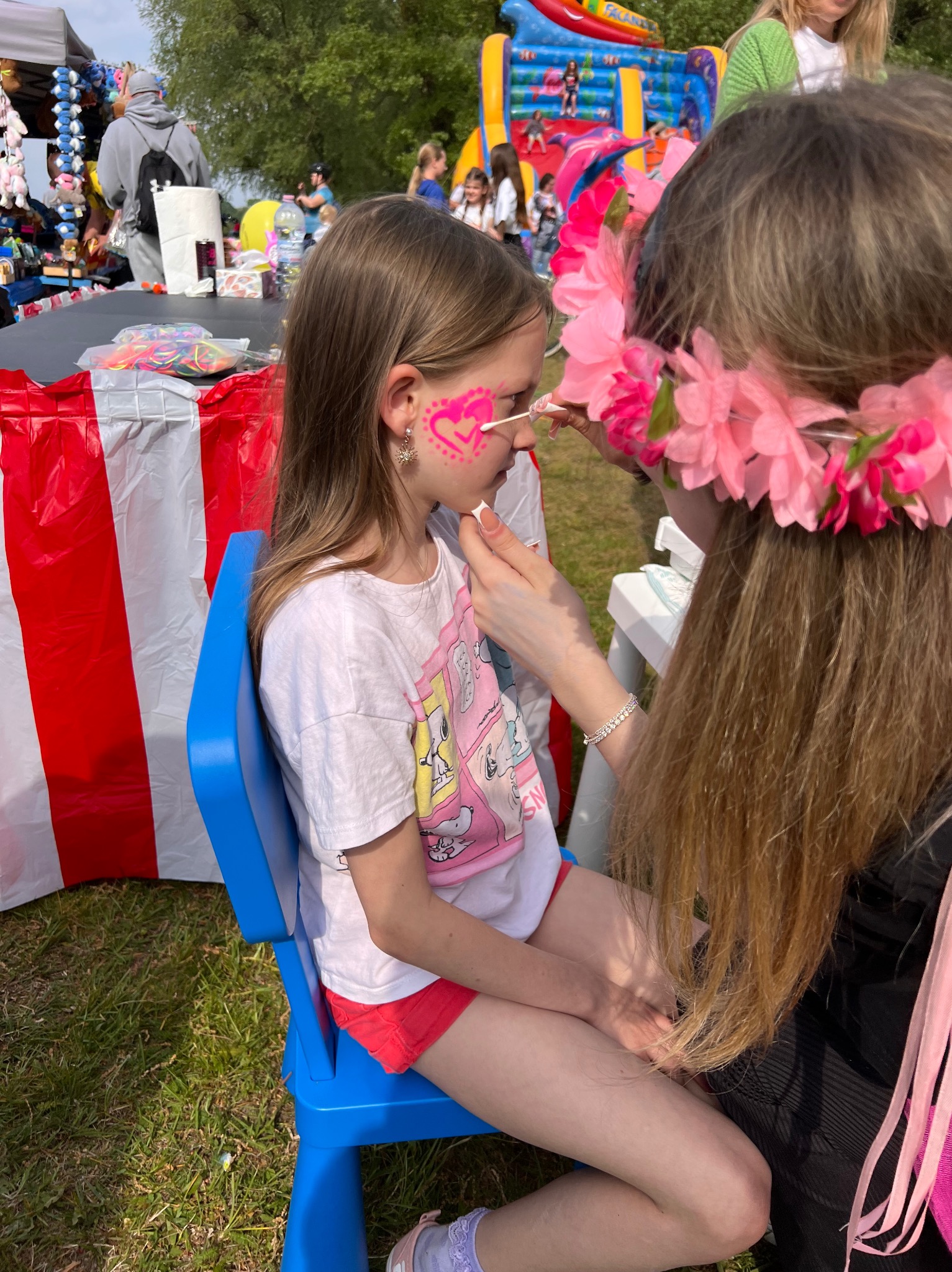 Dziewczynka z różowym sercem malowanym na policzku siedzi na niebieskim krześle. Artystka w wianku z kwiatów maluje jej twarz. W tle dmuchany zamek.
