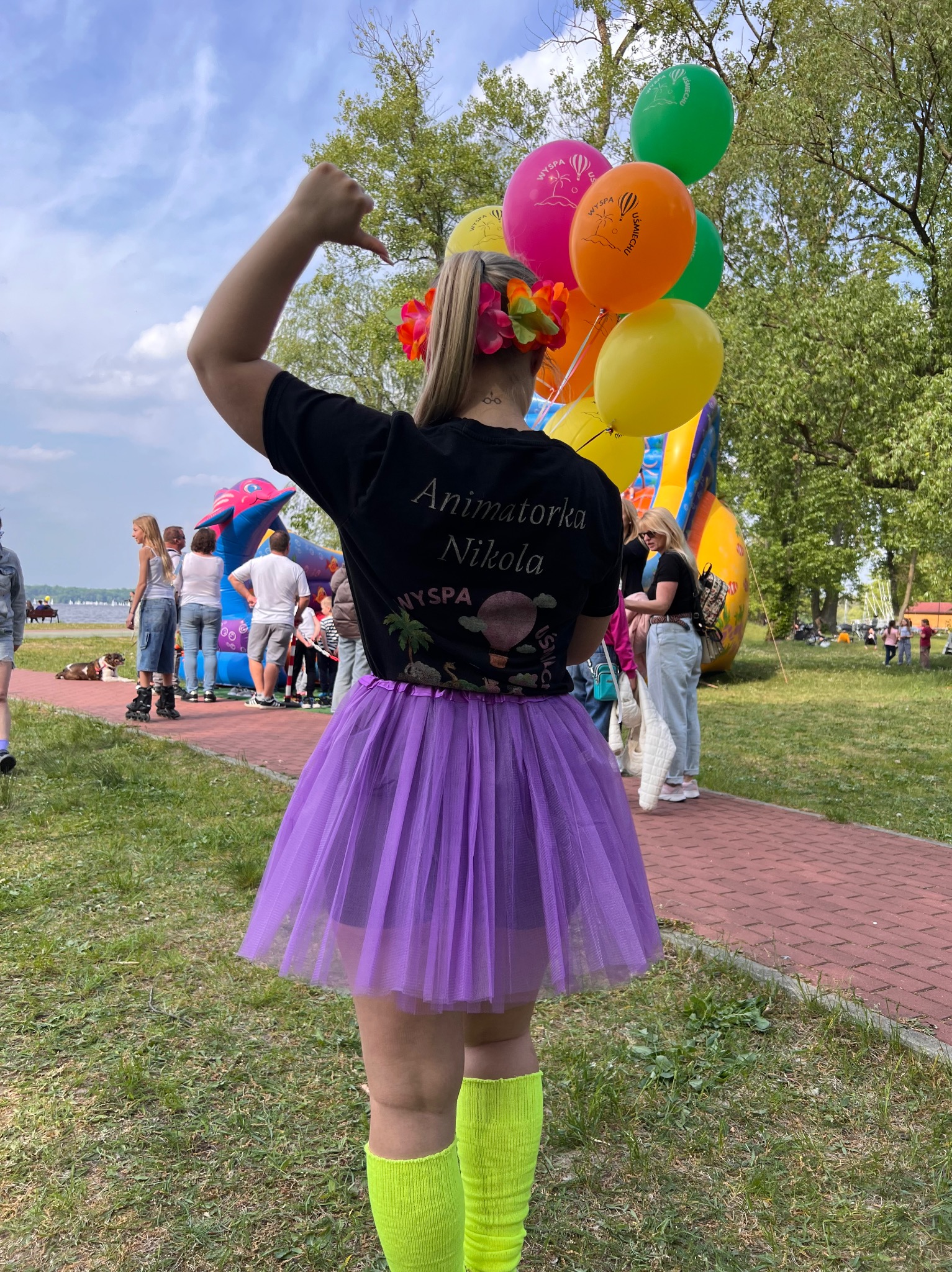 Kobieta w czarnej koszulce z napisem 'Animatorka Nikola', fioletowej spódnicy tutu, trzymająca balony na tle dmuchanego zamku i ludzi na imprezie plenerowej.