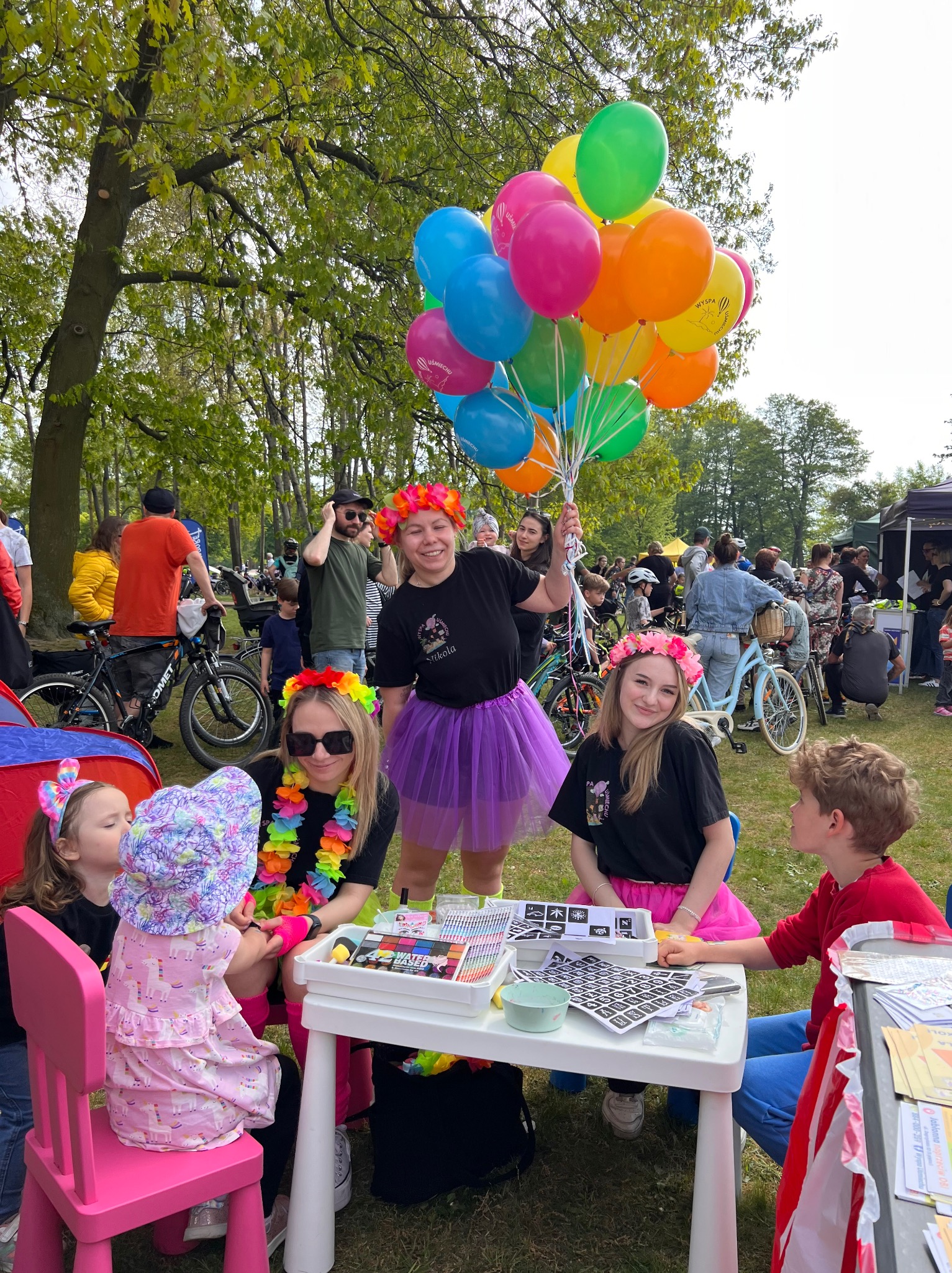 Animatorzy dla dzieci, w kolorowych strojach, prowadzą warsztaty plastyczne na świeżym powietrzu. Dzieci siedzą przy białym stole, otoczone balonami i rekwizytami.