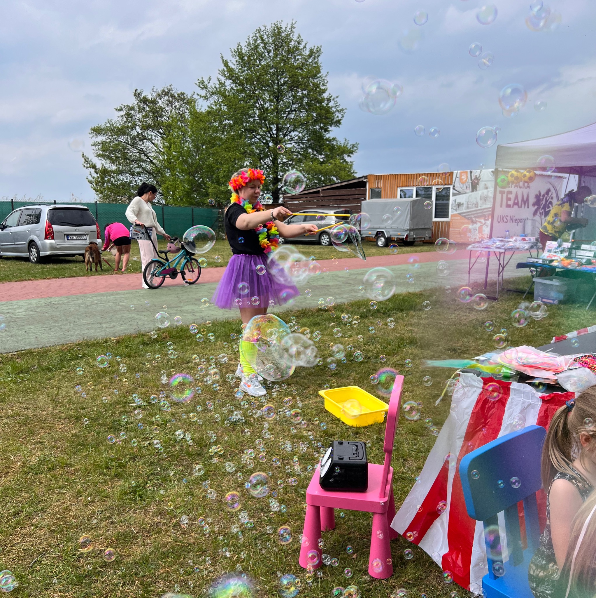 Animator w hawajskim stroju puszcza bańki mydlane na trawie. W tle dzieci, samochód i przyczepa. Kolorowe rekwizyty i dużo baniek tworzą radosną atmosferę.