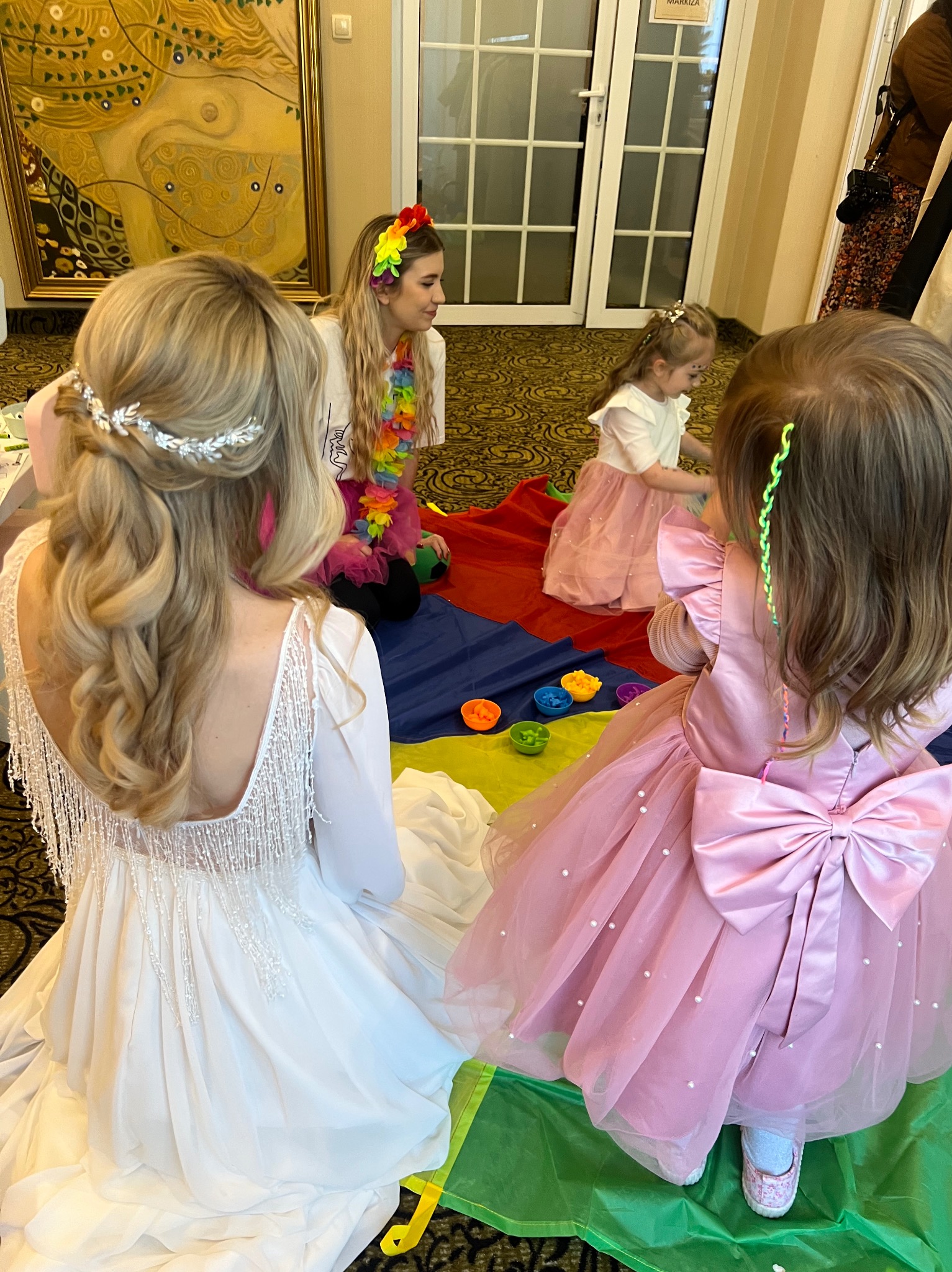 Animator w tęczowej girlandzie i dwie dziewczynki w sukienkach bawią się na kolorowej chuście animacyjnej podczas imprezy dla dzieci w sali z obrazem.