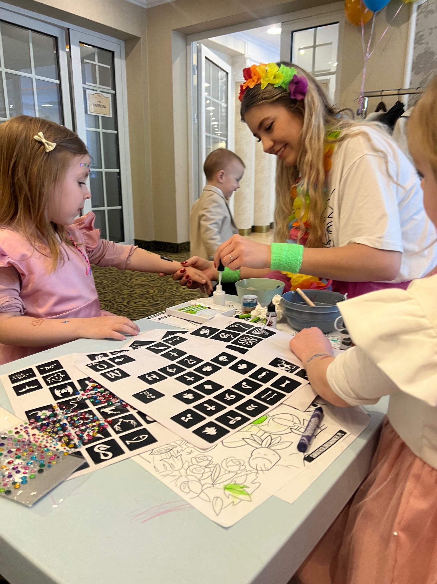 Animator w stroju, z tęczową opaską, maluje brokatowy tatuaż na ręce dziewczynki. Na stole szablony, farbki i kolorowe brokaty. Sala z oknami w tle.