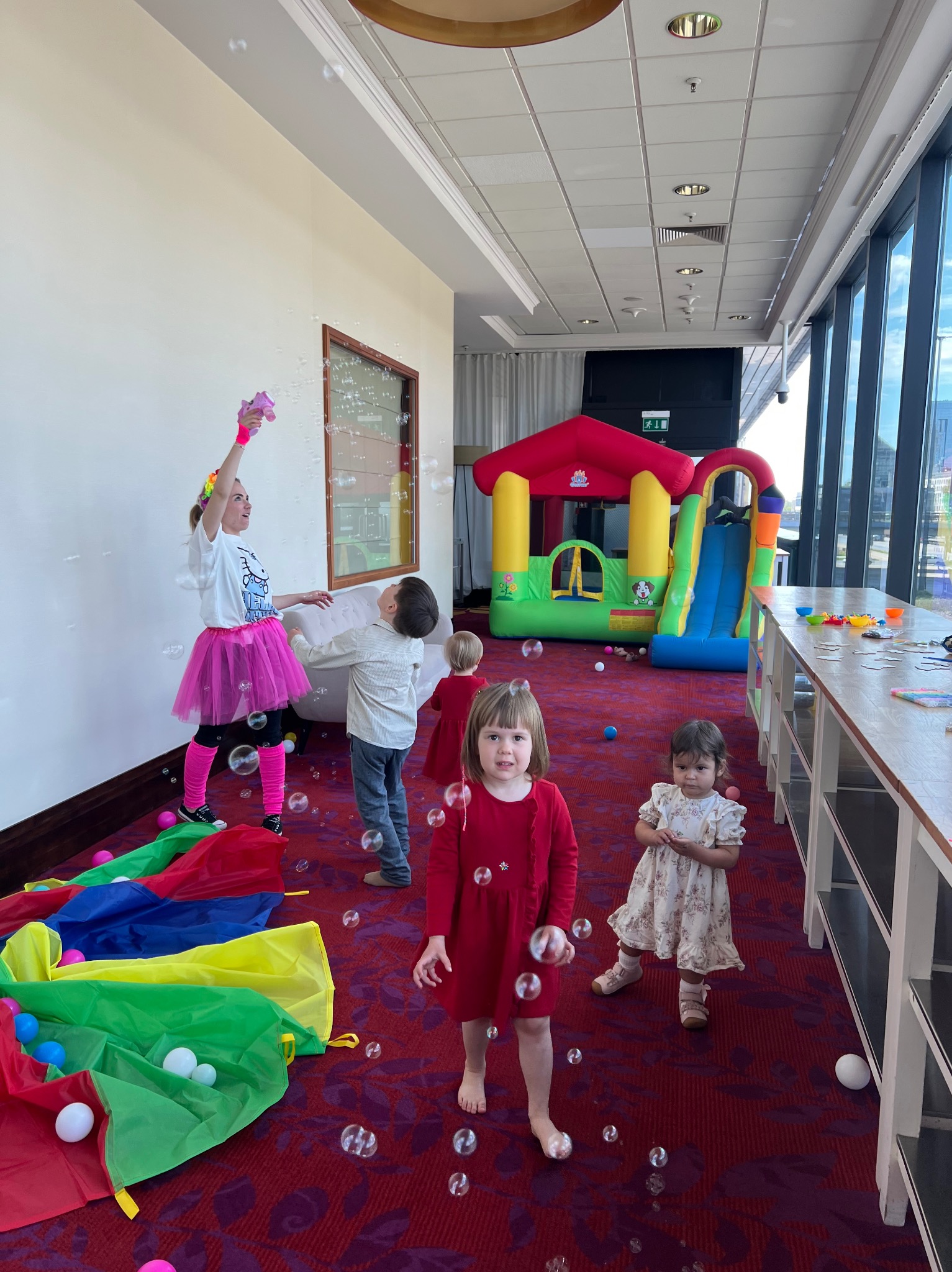 Animator w różowej spódniczce puszcza bańki mydlane podczas zabawy z dziećmi, w tle dmuchany zamek, kolorowa chusta animacyjna i stół z akcesoriami.