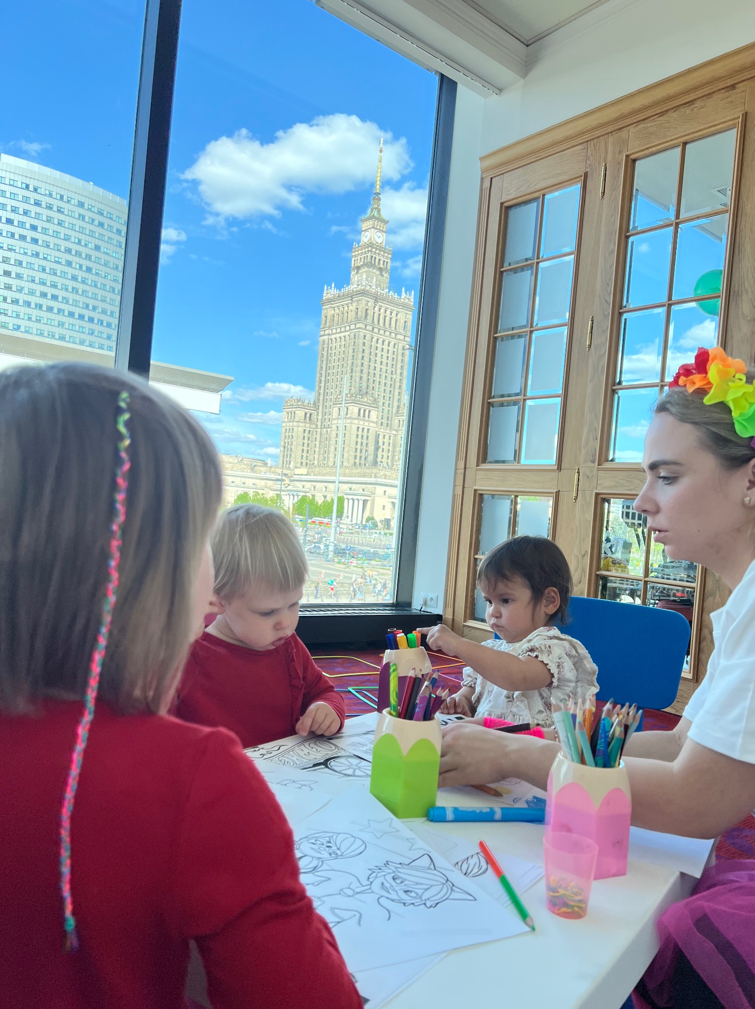 Animator z dziećmi koloruje kolorowanki przy stole, w tle Pałac Kultury i Nauki w Warszawie, widok z okna. Kolorowe kredki i pisaki na stole.