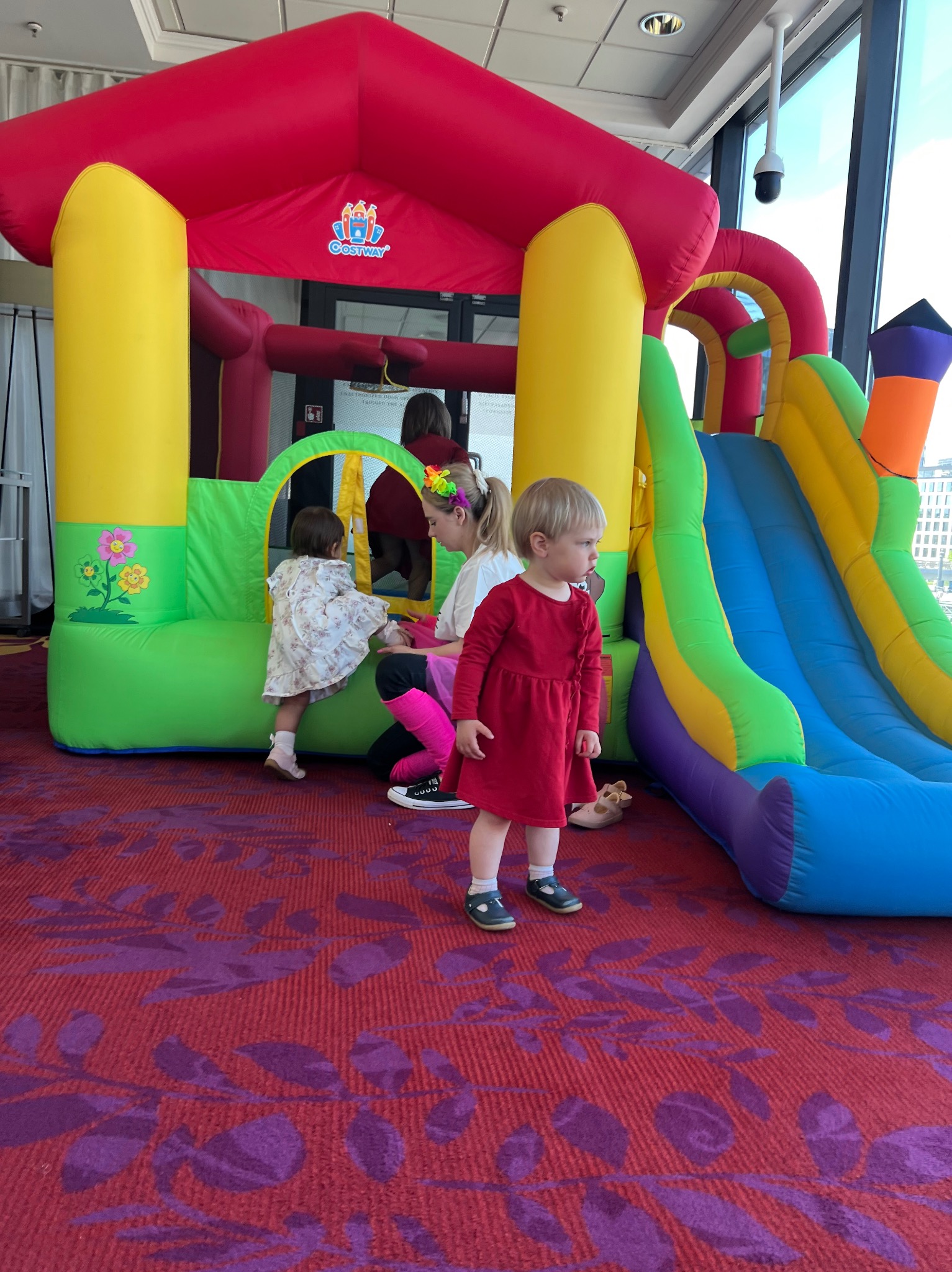 Dziewczynka w czerwonej sukience stoi przed dmuchanym zamkiem z kolorową zjeżdżalnią. W tle widać animatorkę z dzieckiem wewnątrz zamku.