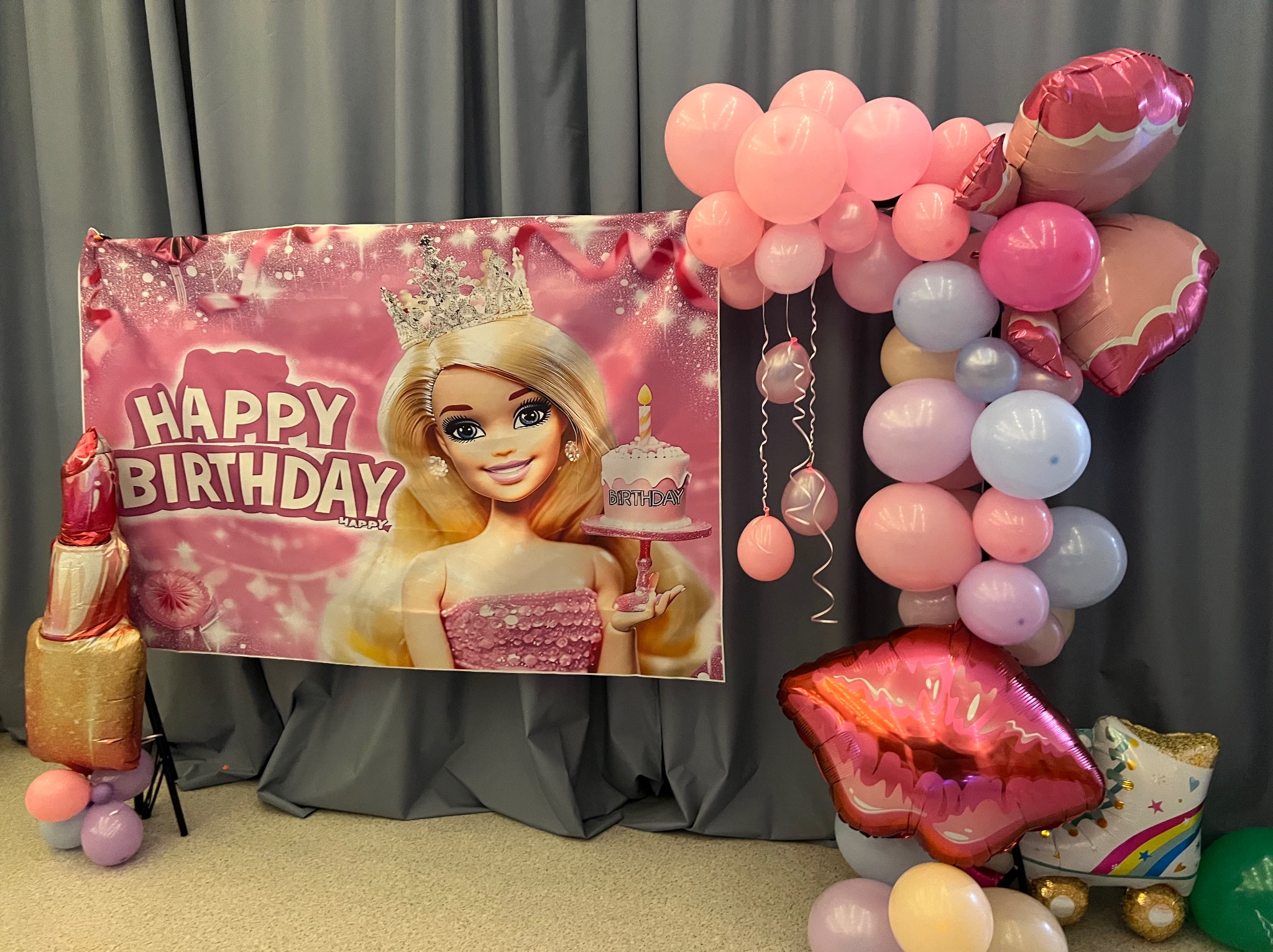 Różowa dekoracja urodzinowa z motywem Barbie: baner z lalką w koronie, balony w odcieniach różu, foliowe balony w kształcie szminki i wrotek.
