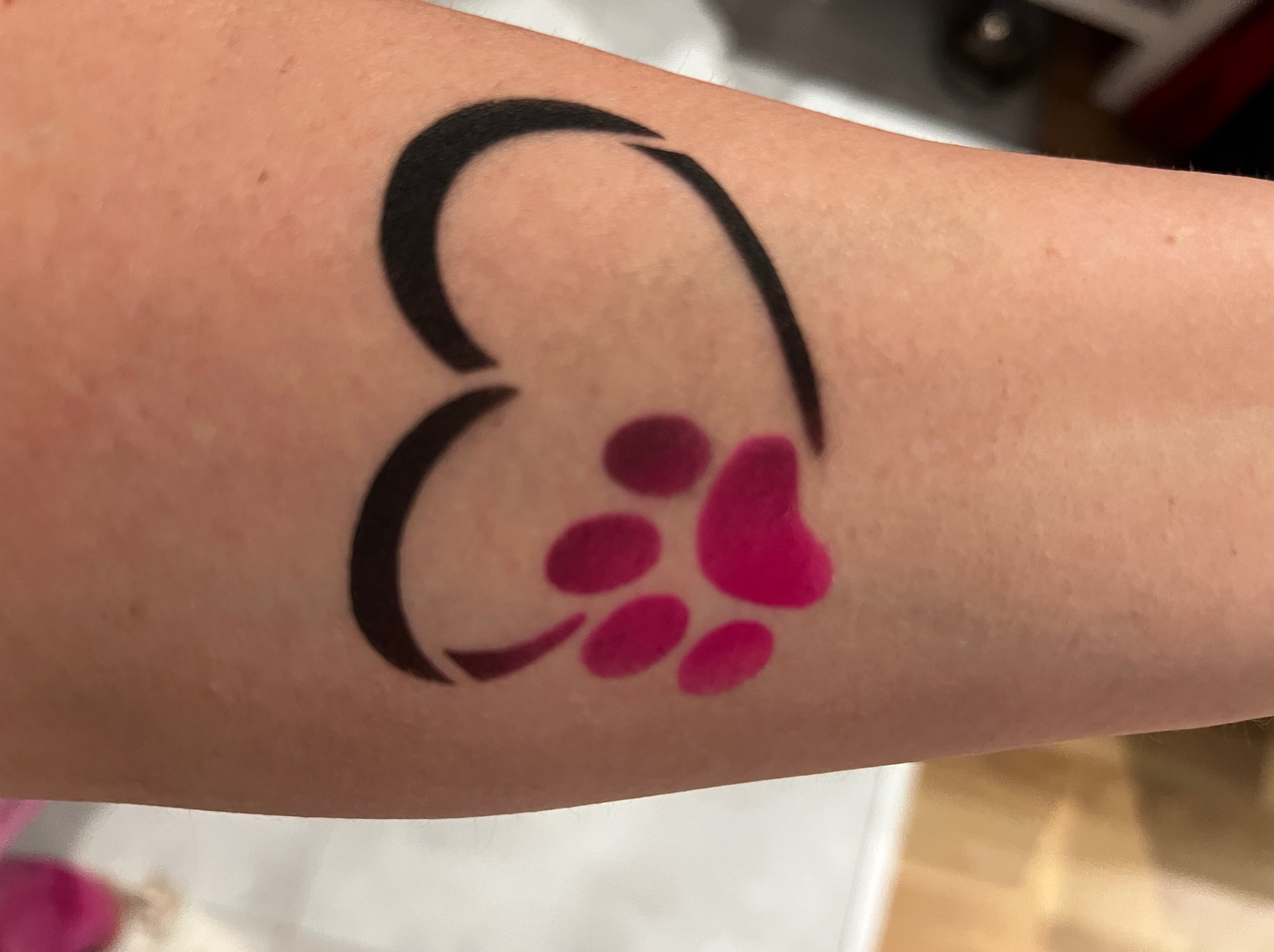 Tatuaż na ramieniu przedstawiający czarny kontur serca z różową łapą zwierzęcia w środku. Widoczne delikatne nierówności skóry.