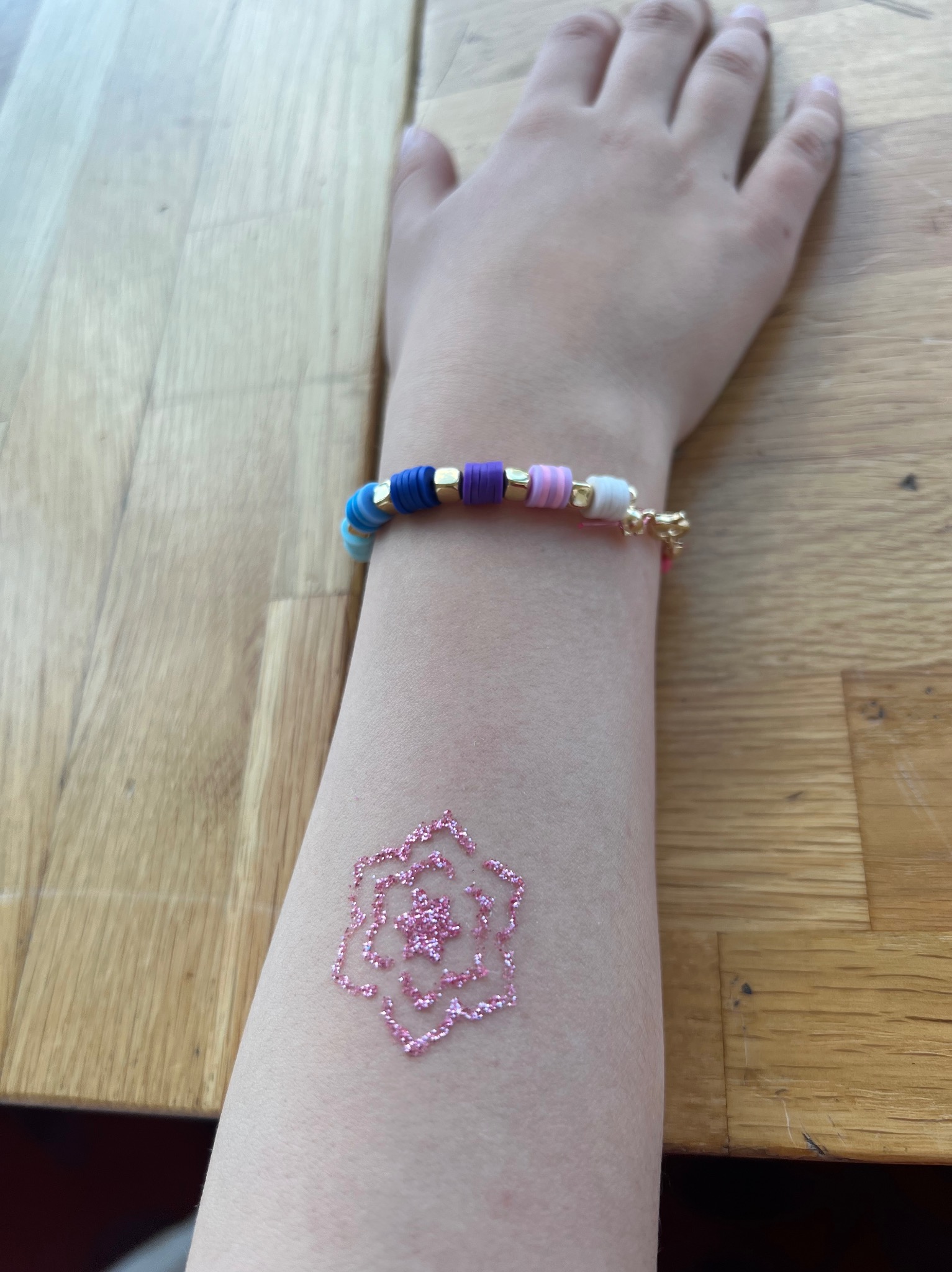 Ramię dziecka z brokatowym tatuażem w kształcie kwiatu i kolorową bransoletką na tle drewnianego stołu. Jasne, naturalne światło.