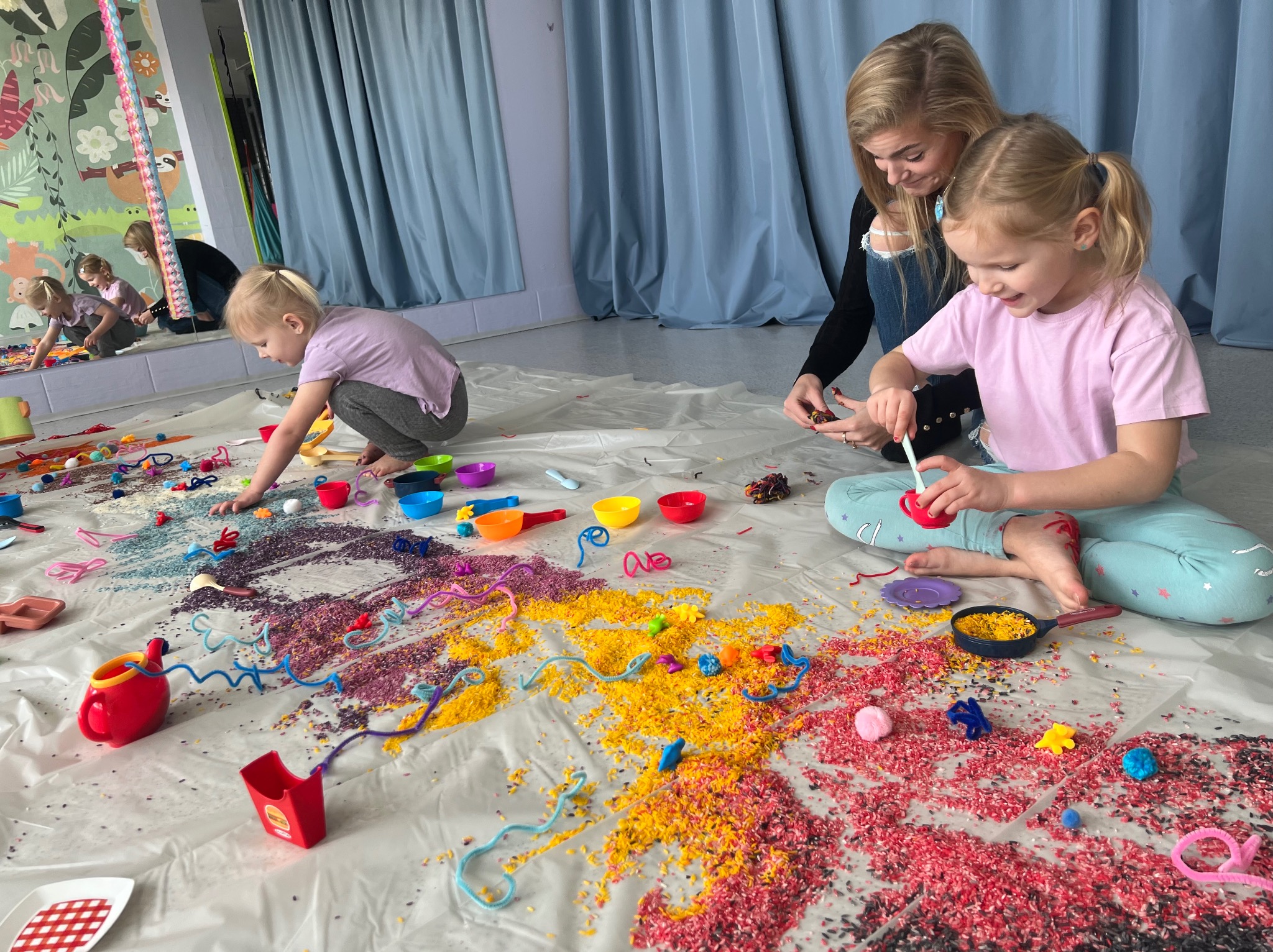 Dwie dziewczynki i kobieta bawią się sensorycznie na podłodze, rozsypane kolorowe ryże, miseczki i zabawki. Lustro w tle odbija bawiące się dzieci.