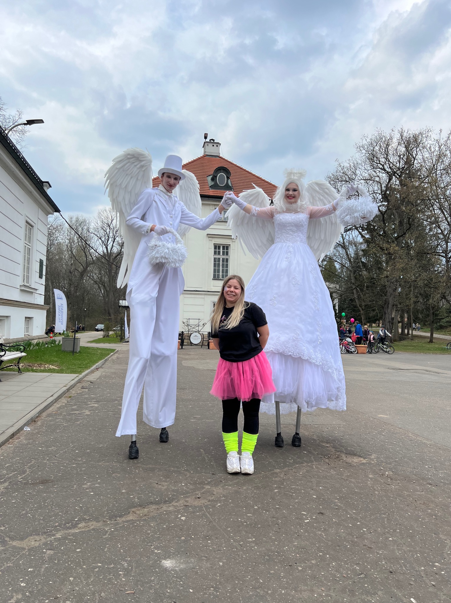 Dwoje animatorów na szczudłach w strojach aniołów pozuje z uśmiechniętą dziewczyną w neonowej spódniczce. W tle park i budynek z czerwoną dachówką.
