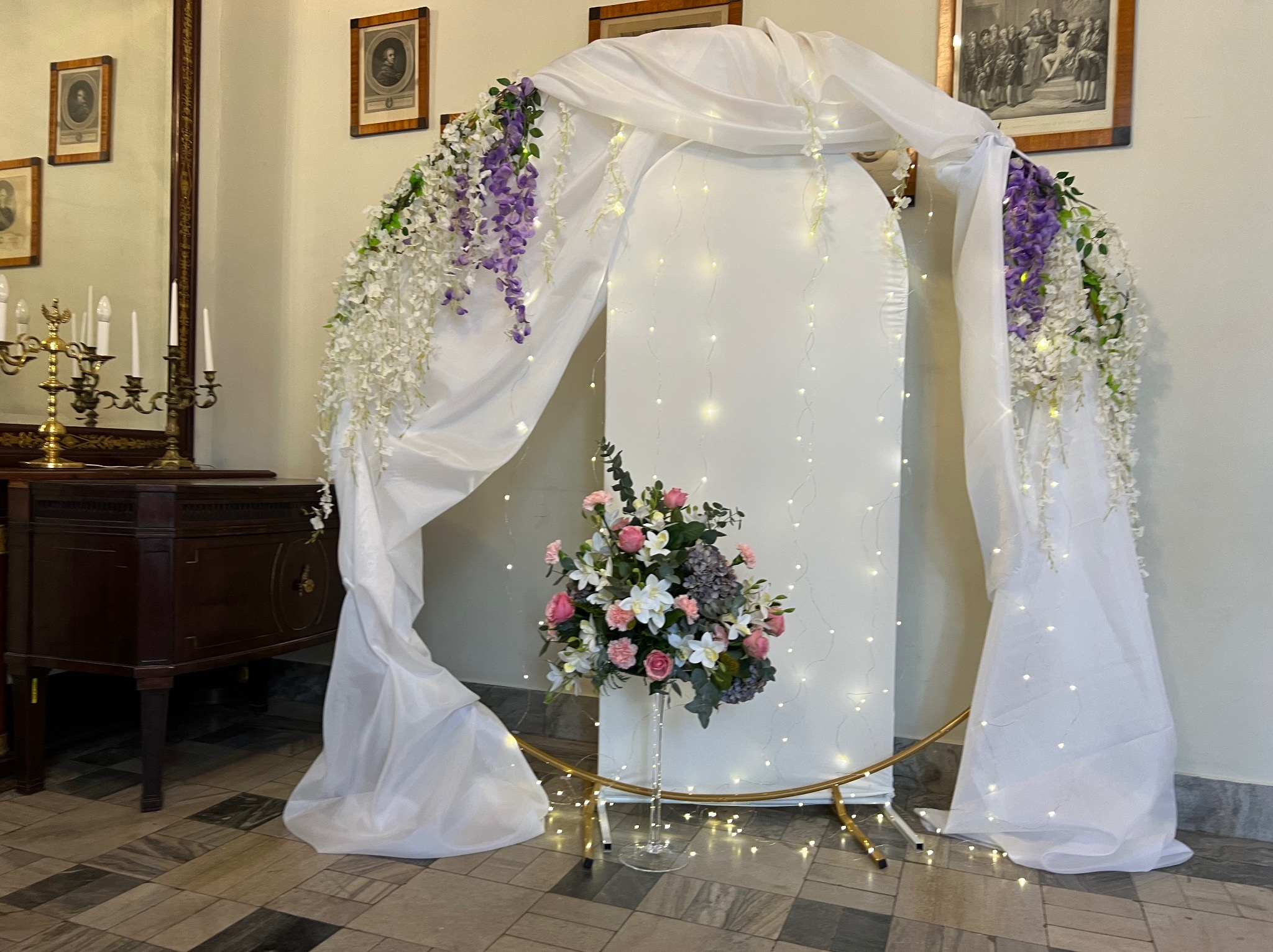 Dekoracja ślubna: biały łuk z kwiatami w odcieniach fioletu i bieli, oświetlony delikatnymi lampkami, z bukietem w szklanym wazonie. Klasyczne wnętrze z obrazami i komodą.