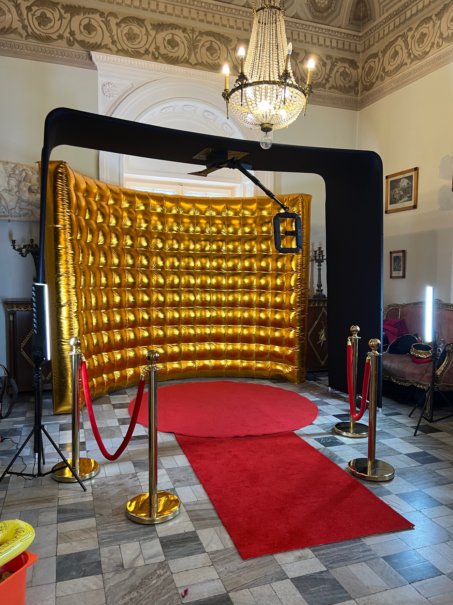 Złota, dmuchana ścianka fotobudki z czarną ramą i czerwonym dywanem w eleganckim wnętrzu z żyrandolem. Gotowa strefa do zdjęć na imprezie.