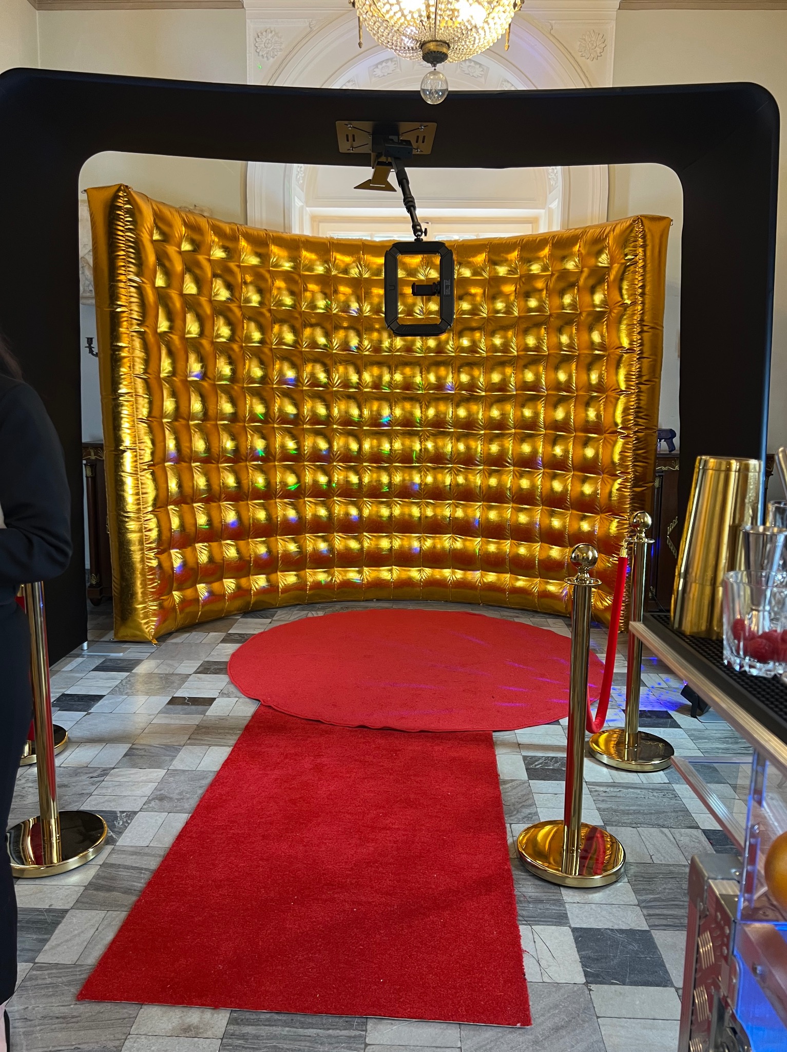 Złota, dmuchana ścianka fotobudki z czarną ramą i czerwonym dywanem. W tle widoczny fragment baru z akcesoriami. Całość w eleganckim wnętrzu.