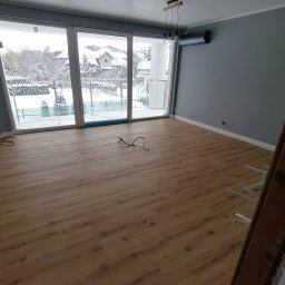 Perfect-bud - Jasne panele podłogowe w nowym pokoju z dużym oknem balkonowym i szarą ścianą. Widoczne kable i drabina. Za oknem zimowy krajobraz.