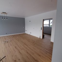 Perfect-bud - Jasne panele podłogowe w przestronnym pokoju po remoncie, szara ściana z lampą. Widoczne przewody elektryczne i gniazdka, przygotowane do wykończenia. Okno w tle.