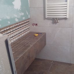Perfect-bud - Łazienka w trakcie wykończenia: brązowe płytki imitujące marmur na podłodze i blacie, częściowo położona mozaika, grzejnik drabinkowy, poziomica i miarka.
