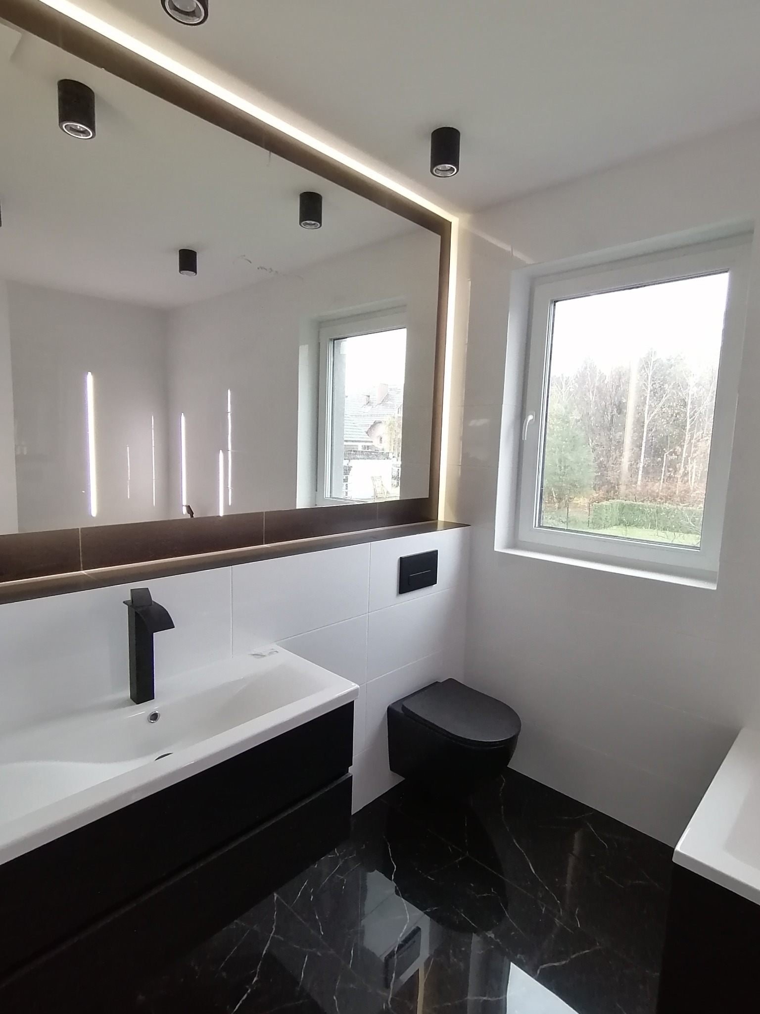 Nowoczesna łazienka z czarnymi akcentami: umywalka, bateria, toaleta. Białe płytki i podświetlane lustro tworzą elegancki kontrast. Widok na zewnątrz przez okno.