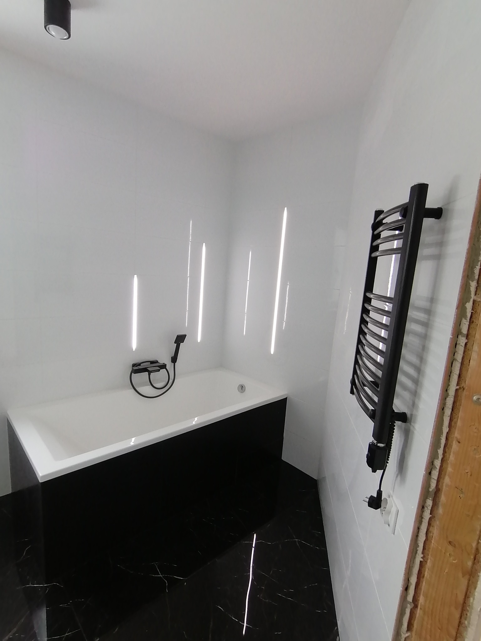 Minimalistyczna łazienka: białe płytki, czarna obudowa wanny z marmurowym wzorem i grzejnik drabinkowy. Widoczne refleksy światła na ścianach.