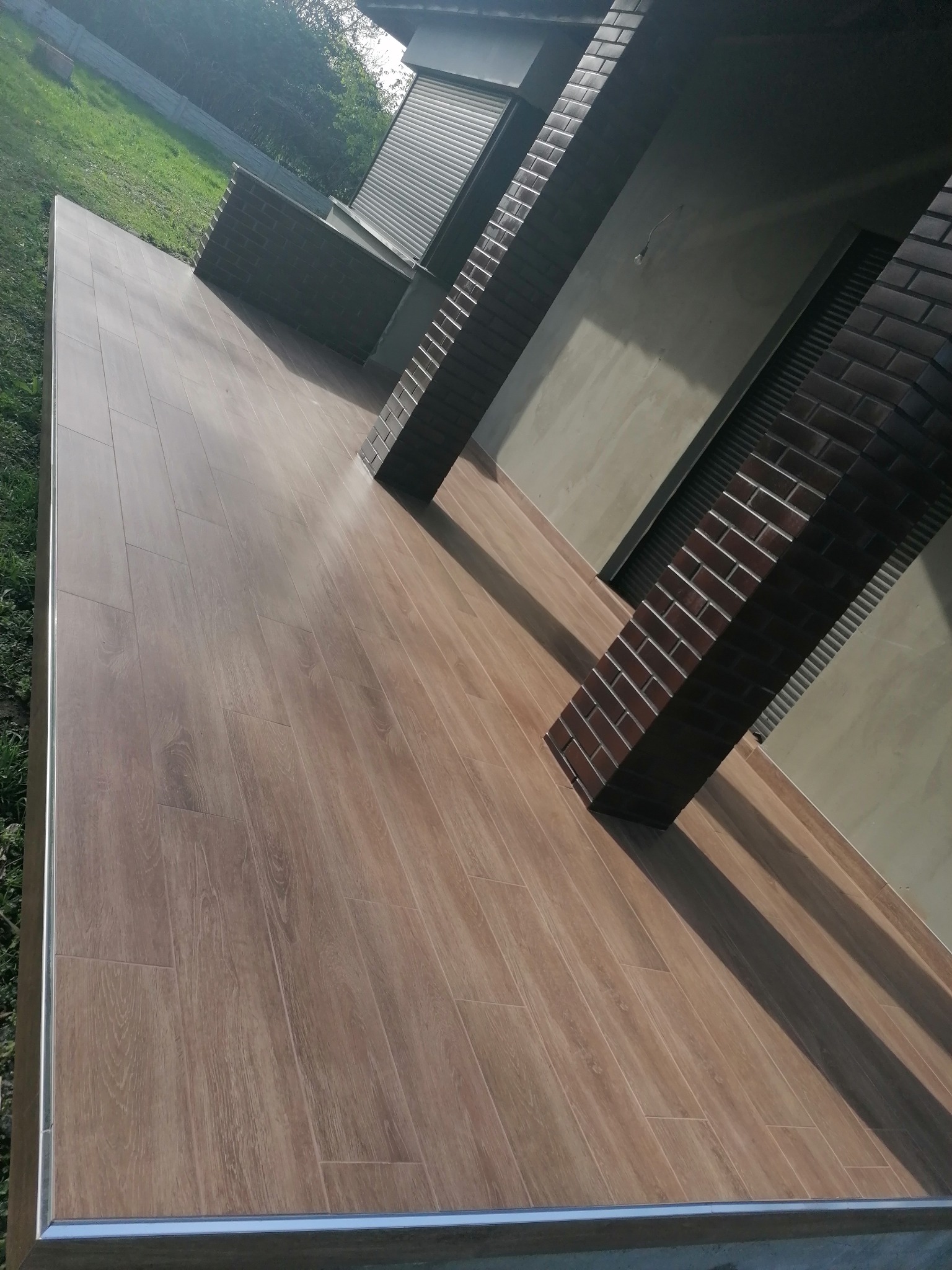 Wykończony taras z drewnopodobną podłogą i ceglanymi filarami. Widoczne detale wykończeniowe, takie jak listwy przypodłogowe. Ujęcie z perspektywy.