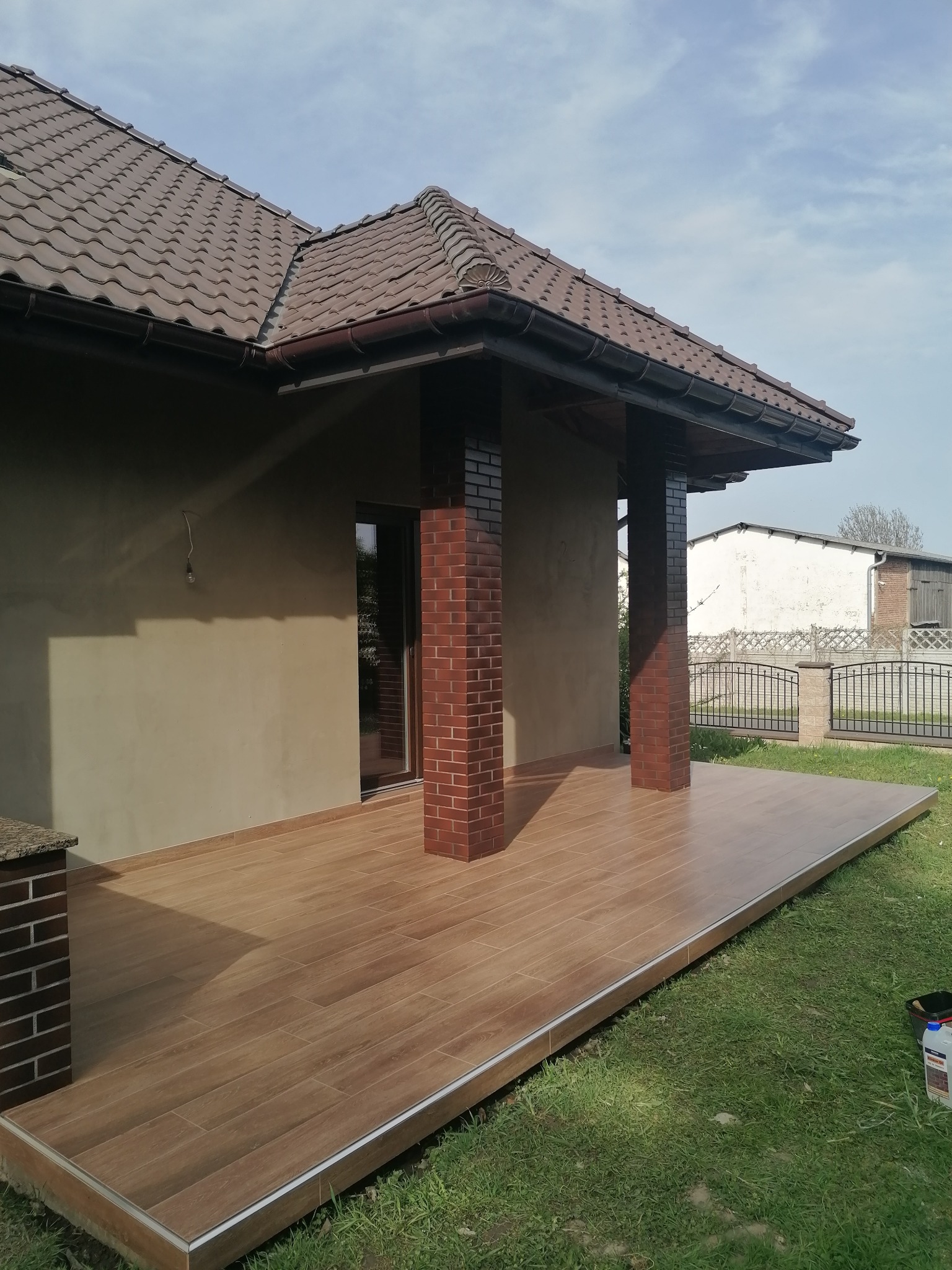 Zadaszony taras z brązową, drewnopodobną terakotą, ceglane filary. Widok na dom z brązowym dachem i zielony trawnik. Częściowo otynkowana elewacja.
